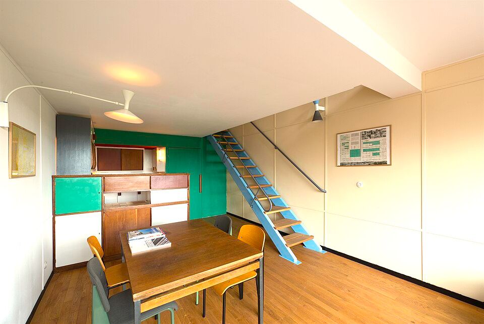Estar y comedor de una vivienda con mesa de madera, armarios de colores y una escalera metálica que sube a un altillo.