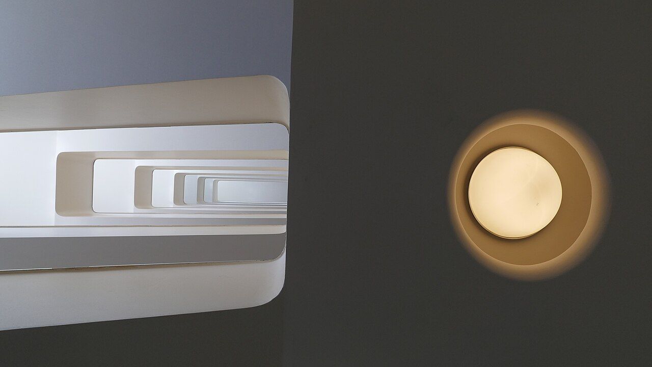 Hueco de una escalera visto desde abajo: barandillas blancas y curvas que se alejan en perspectiva junto a una lámpara redonda.