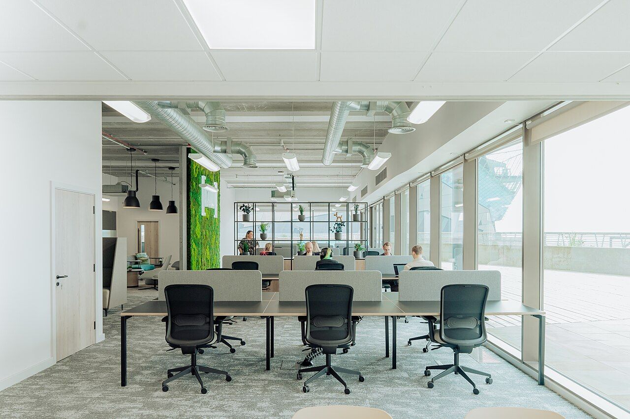 Sala de trabajo diáfana con mesas compartidas, conductos de ventilación vistos en el techo y una cristalera hacia una terraza.