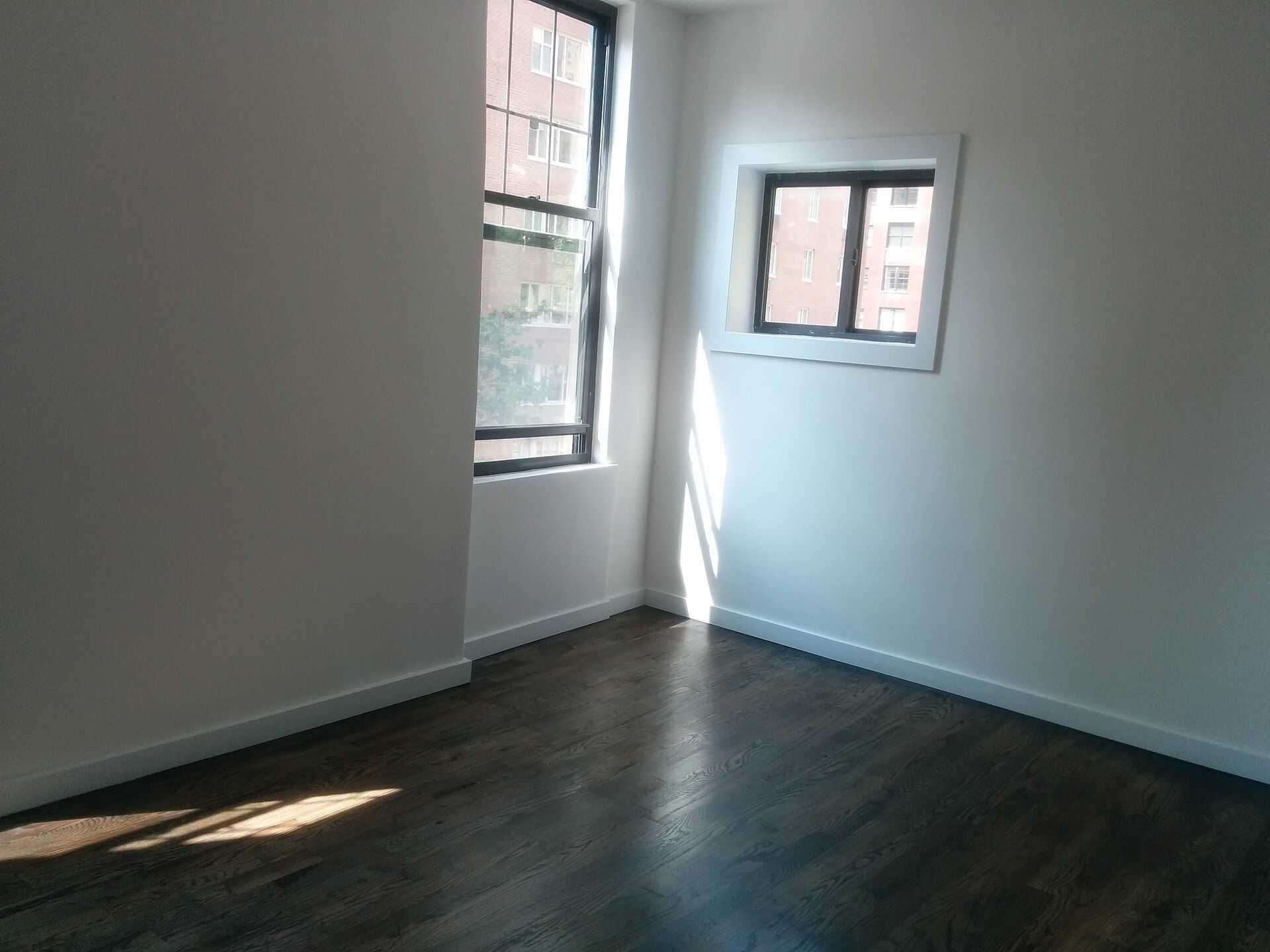 Habitación vacía con suelo de madera oscura, paredes blancas y dos ventanas por las que entra el sol.